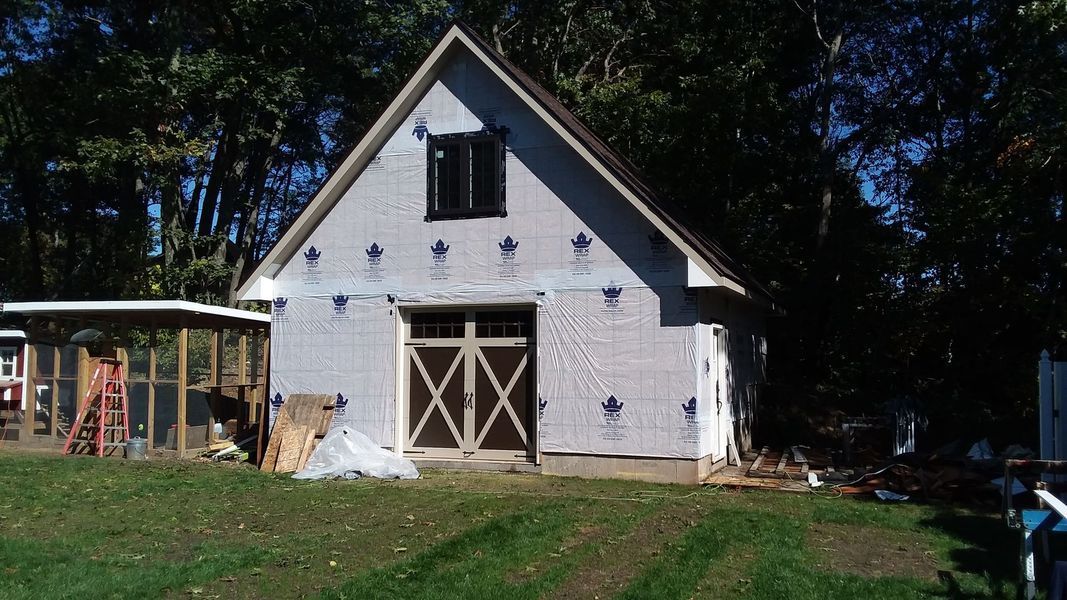 Barn-like structure under construction, with a large garage door, a small window, and building wrap on the exterior.
