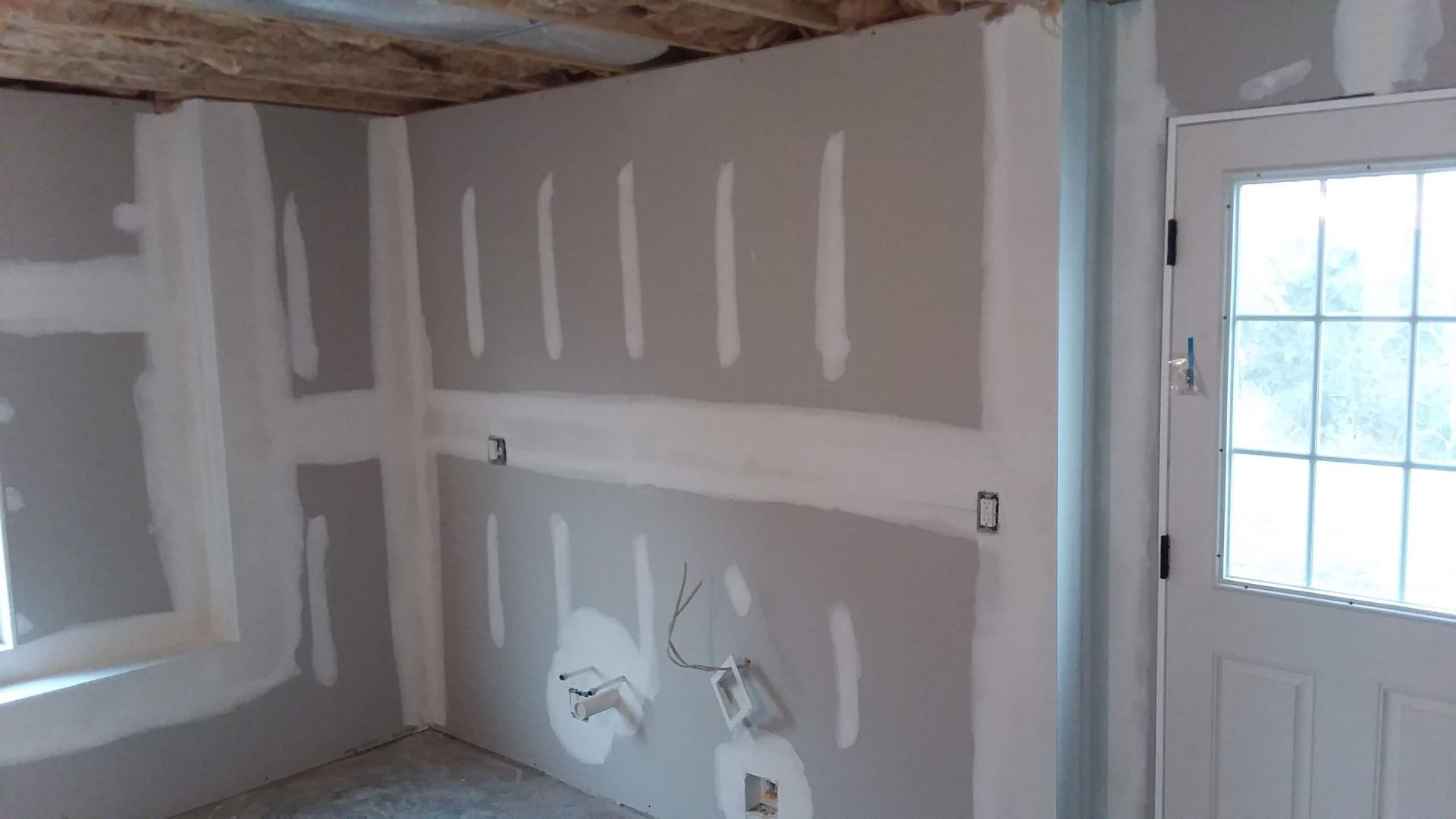 Interior room under construction, with drywall installed, taping and mudding in progress. Door and window are present.