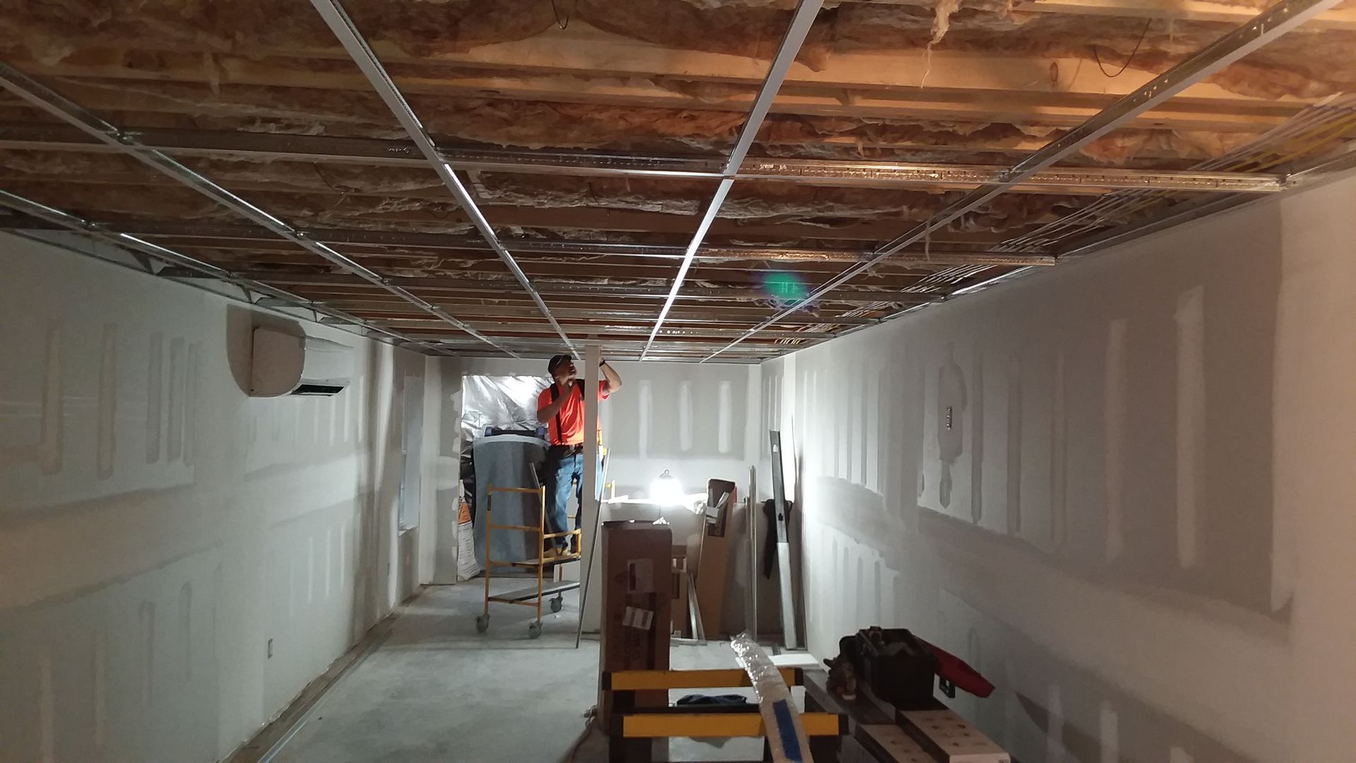 Person on ladder installing ceiling grid in a room with drywall walls.