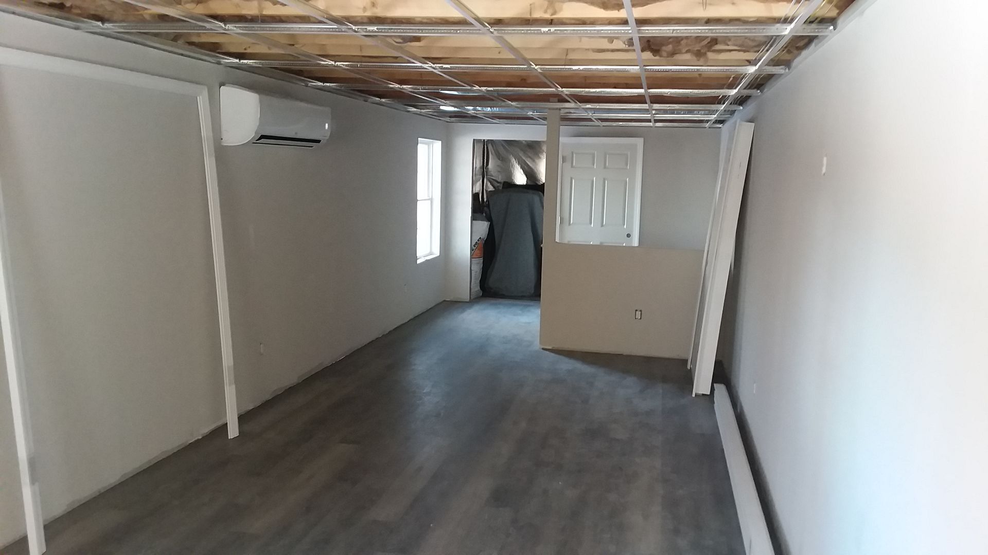 Interior of a room under renovation: white walls, gray flooring, exposed ceiling beams, a door, and a window.