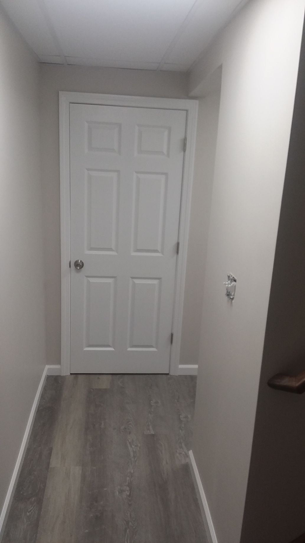Narrow hallway with a closed white door at the end, gray flooring, and light gray walls.