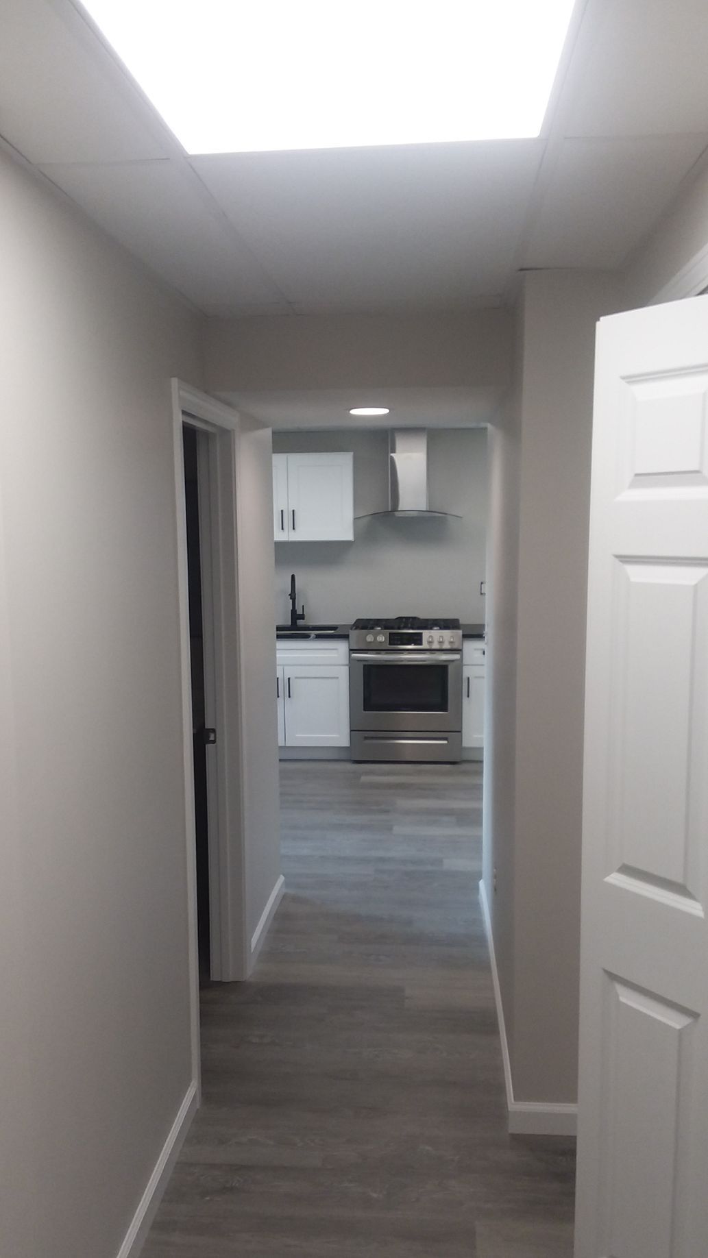 Hallway leading to a kitchen with white cabinets, stainless steel appliances, and grey flooring.