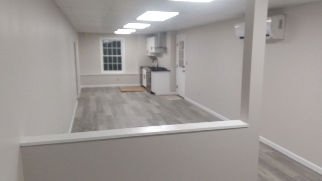 Empty interior with light gray walls, wood-look floor, partial wall and a kitchen at the end.