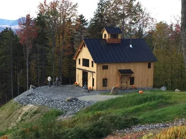 Wooden barn with black roof on a hill, surrounded by trees with fall foliage.