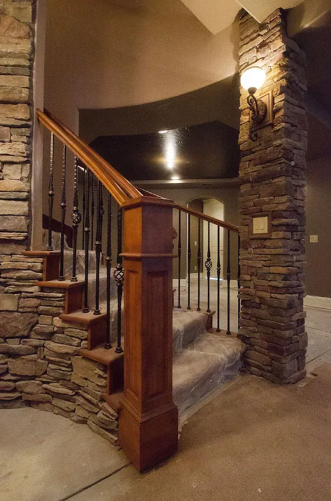 Stone stairwell with wooden handrail, black spindles, and stone columns; indoor setting.