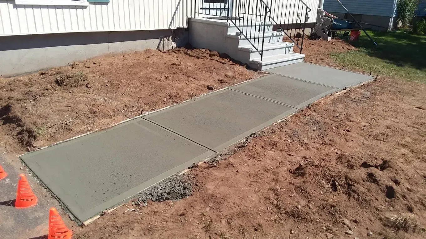 Newly poured concrete walkway leading to a house entrance. Earth-toned ground, orange cones on the left.
