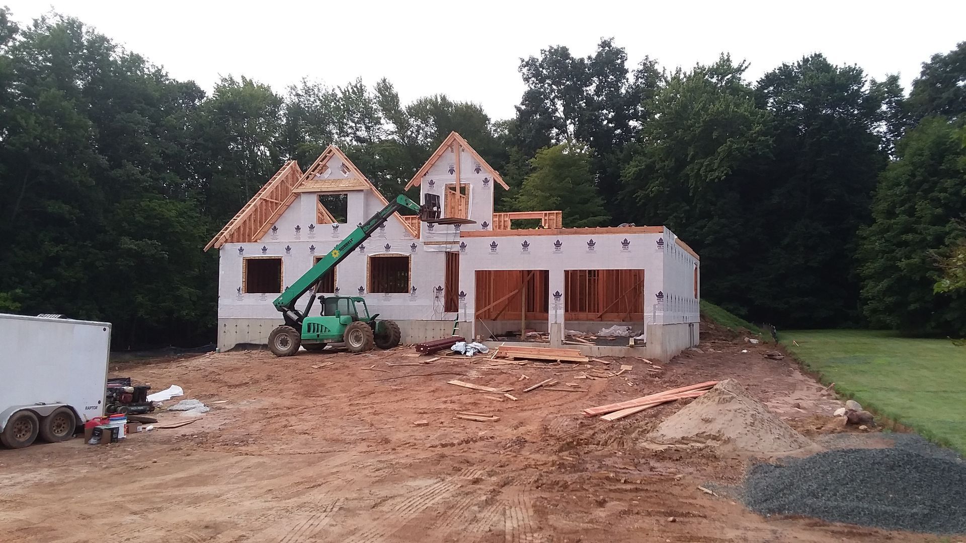 House under construction; framed walls, roof trusses, green lift, dirt and gravel.