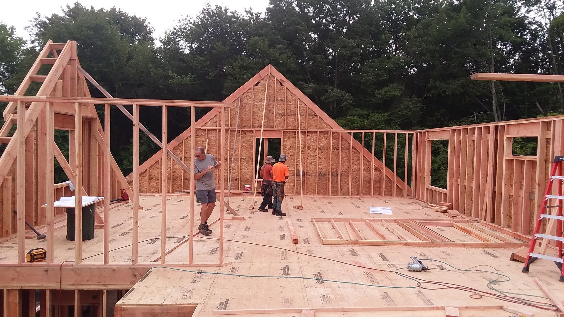Construction workers framing a house with wood; daylight, trees in background.