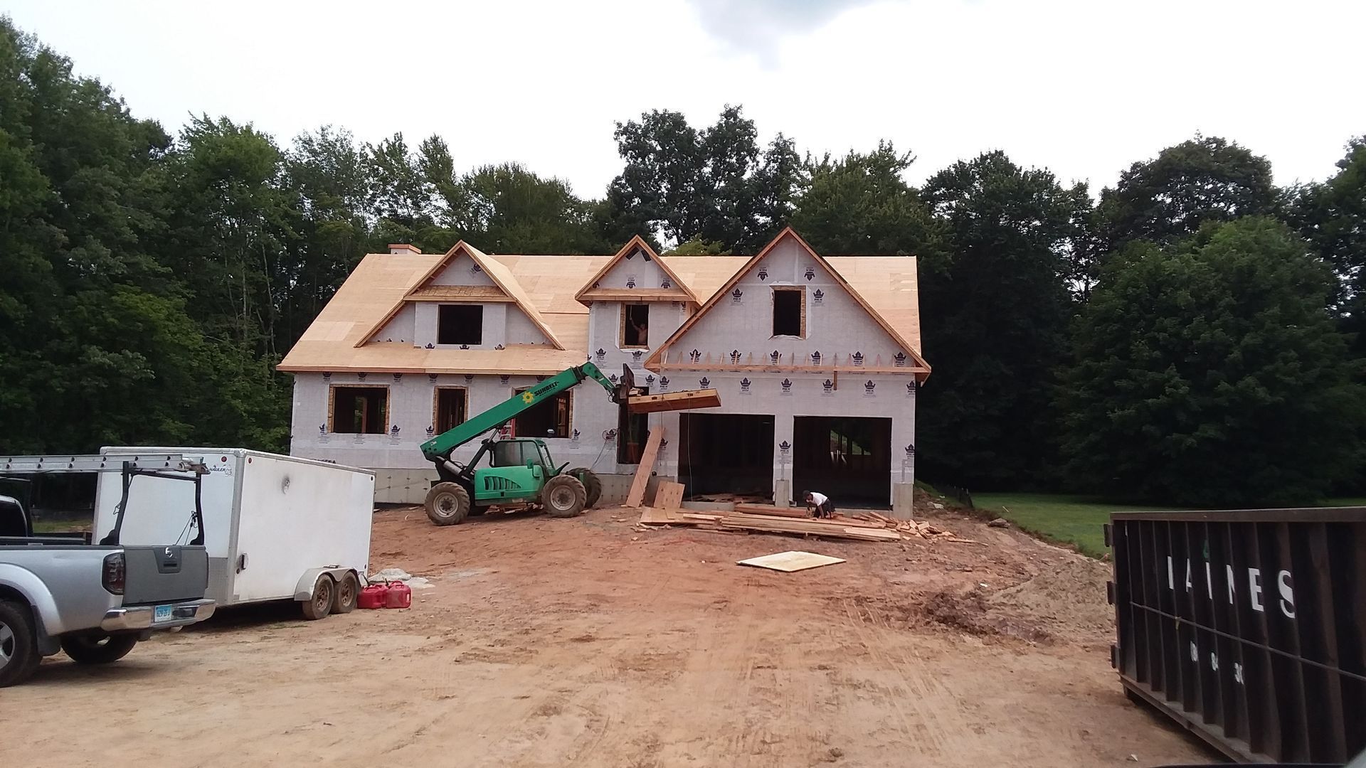 House under construction; workers and machinery on a dirt lot; trees in the background.