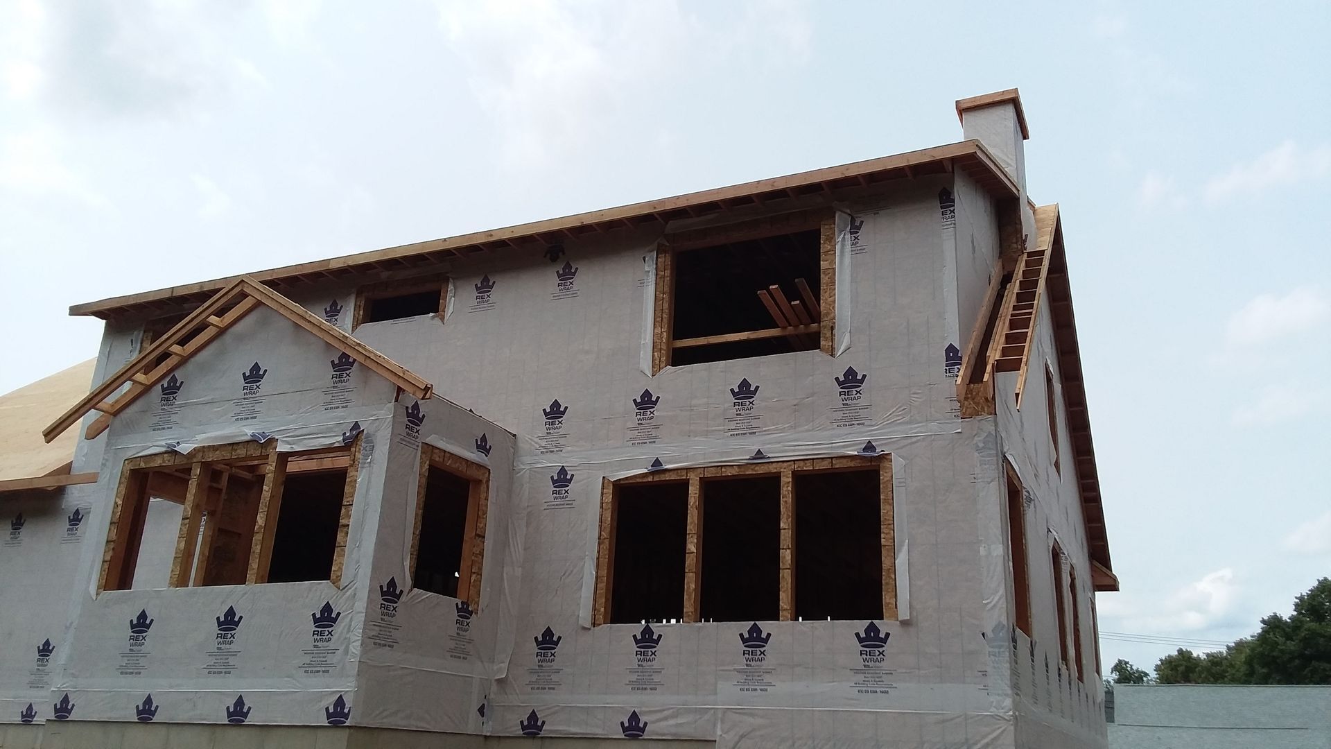 Two-story house under construction, framed with wood, sheathed in grey, with window openings.