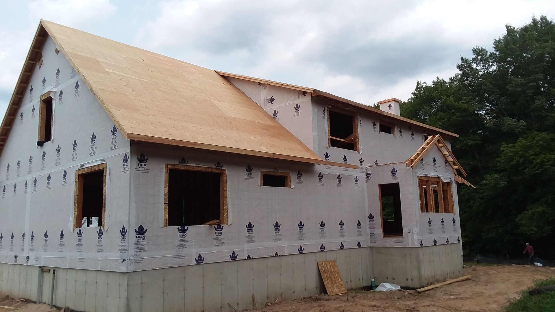 House under construction, sheathed in white, with exposed roof rafters and window frames, set against a cloudy sky.