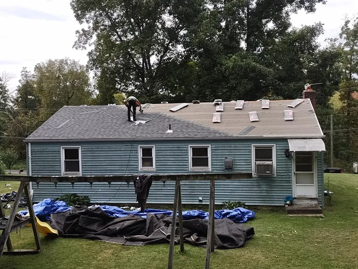 A person roofing a light blue house; tools and tarps sit on the lawn.