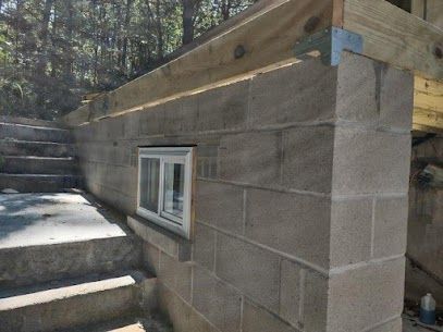 Gray cinder block wall with a white window and wooden beams, next to stone steps.
