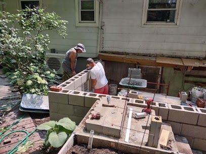 Two people building a cinder block structure next to a house. One person is using mortar.