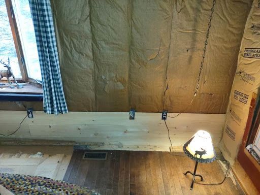 Interior of a cabin with exposed insulation on the angled ceiling, wooden floor, and a lit lamp.