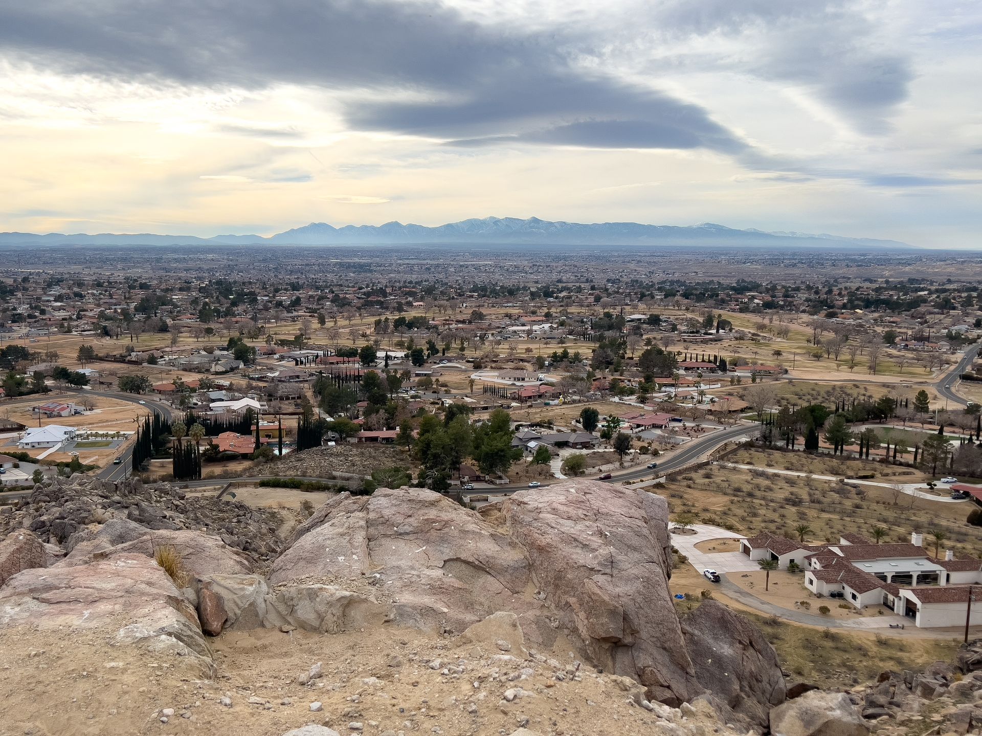 Panoramic view of a city from a rocky hilltop under a cloudy sky. Distant mountains and buildings are visible.