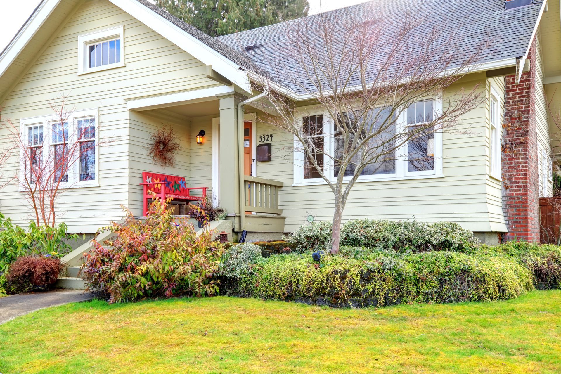 Cottage-style house with green siding, porch, red brick chimney, and landscaping with a small tree and bushes.