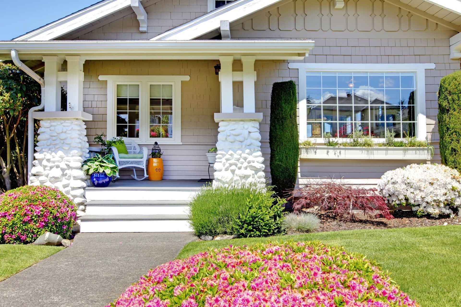 Tan craftsman-style house with porch, garden, and walkway. Pink and green landscaping.