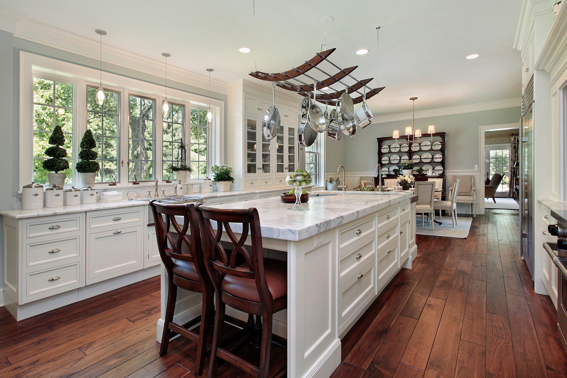 Spacious kitchen with a white island, pot rack, and hardwood floors. Windows and a dining area are visible.