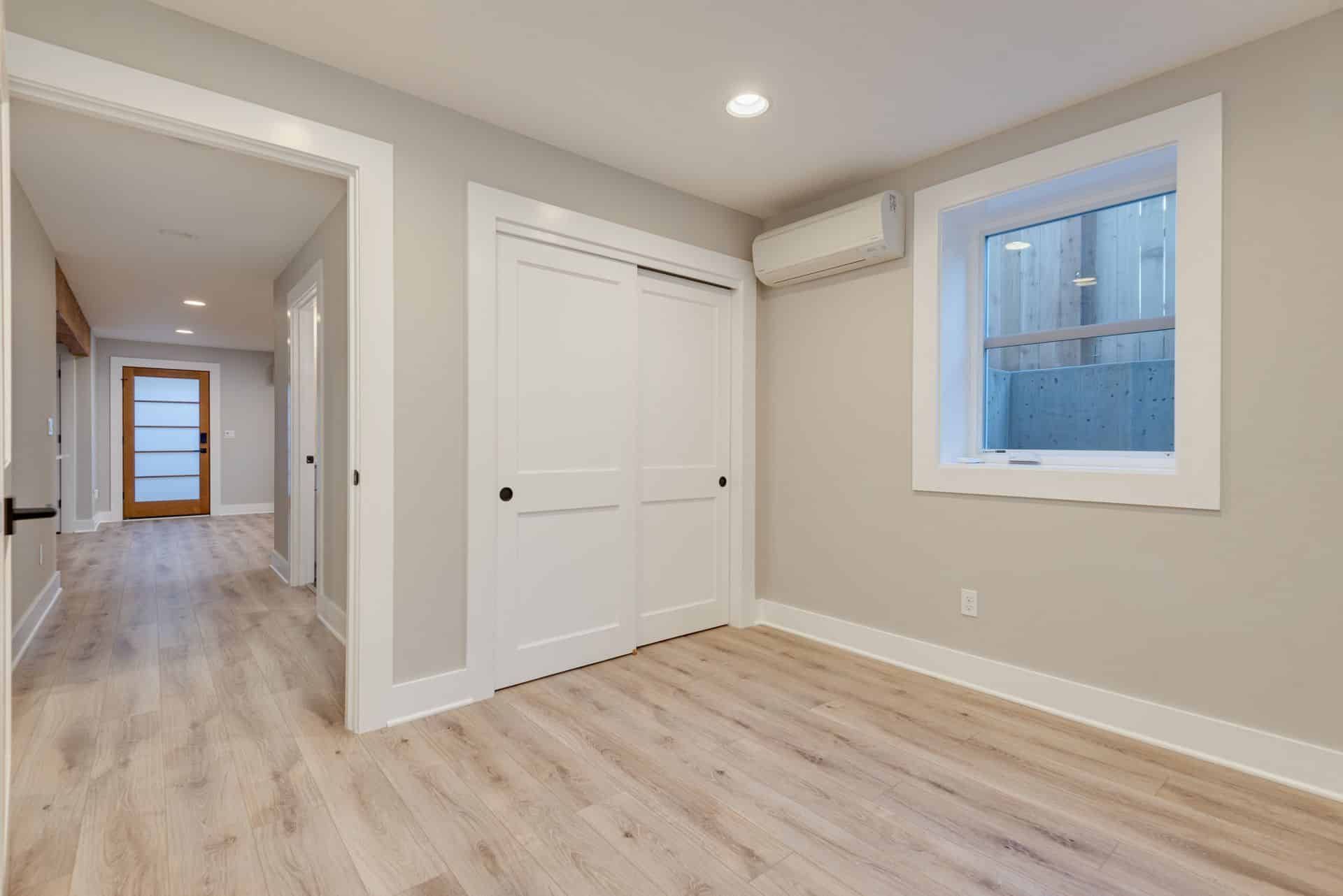 Empty bedroom with wood flooring, white sliding closet doors, and a small window.