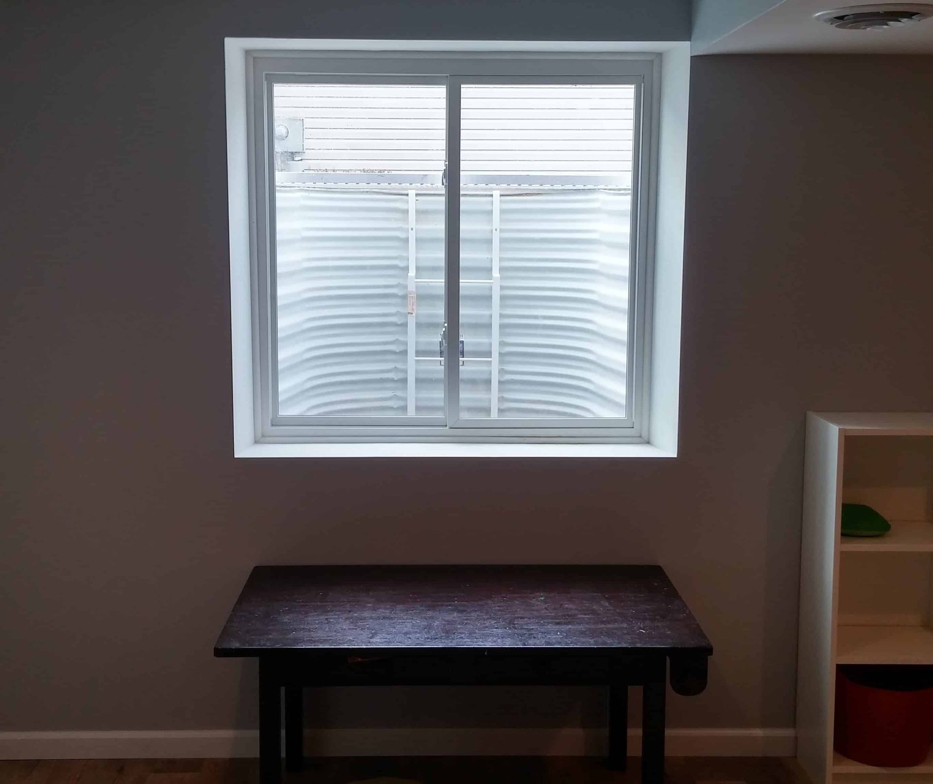 Window in a basement with a small table below it. A white shelf is to the right.