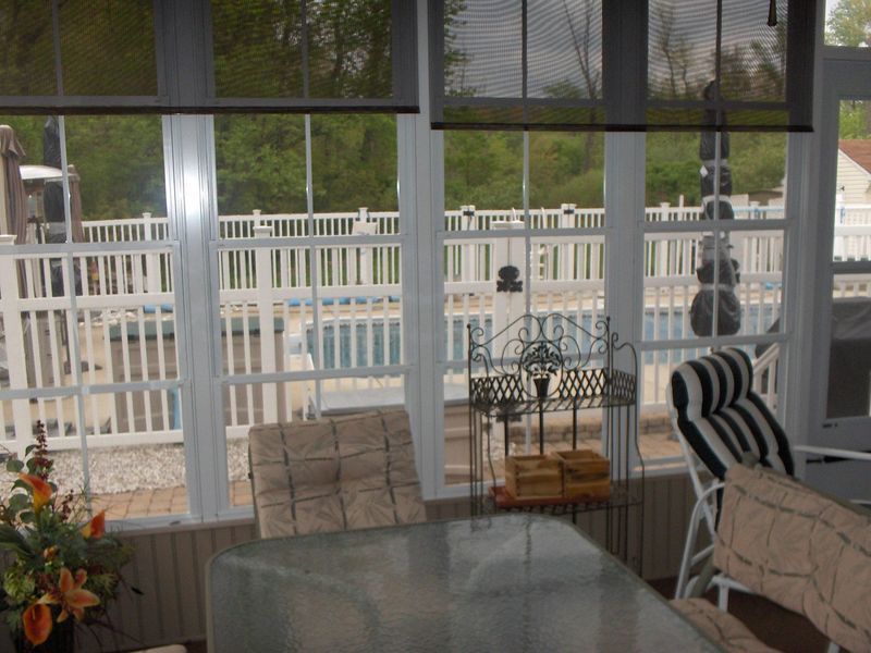Sunroom interior with view of a pool. White-framed windows, roll-up shades. Outdoor furniture.