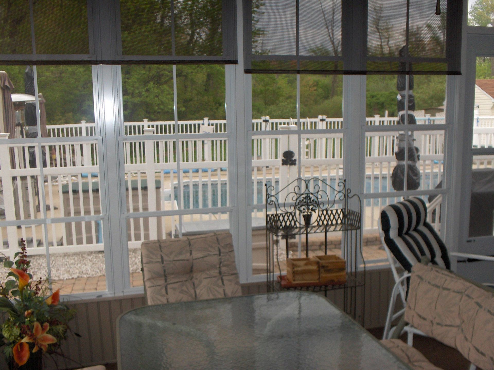 Sunroom with pool view; glass table, chairs, white fence, and greenery outside.