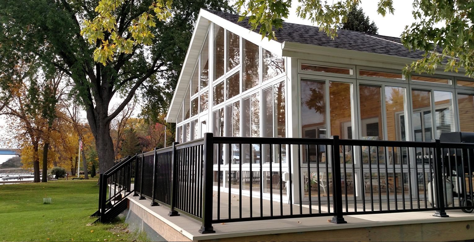 A sunroom with a black fence, on a stone base, next to a grassy area and a tree.
