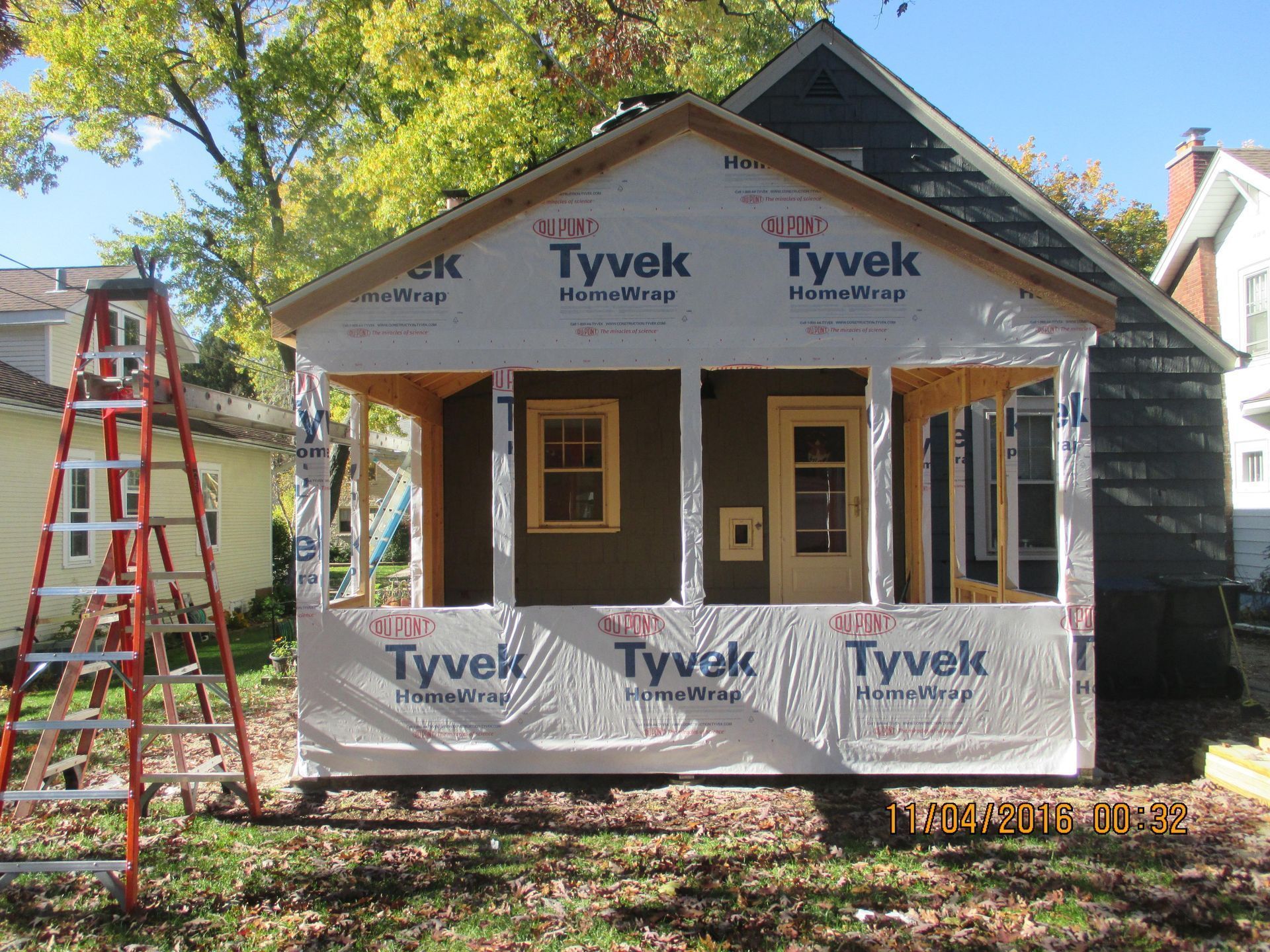 House under construction; Tyvek wrap, porch with brown siding and tan trim, ladder on the left, foliage, sunny day.