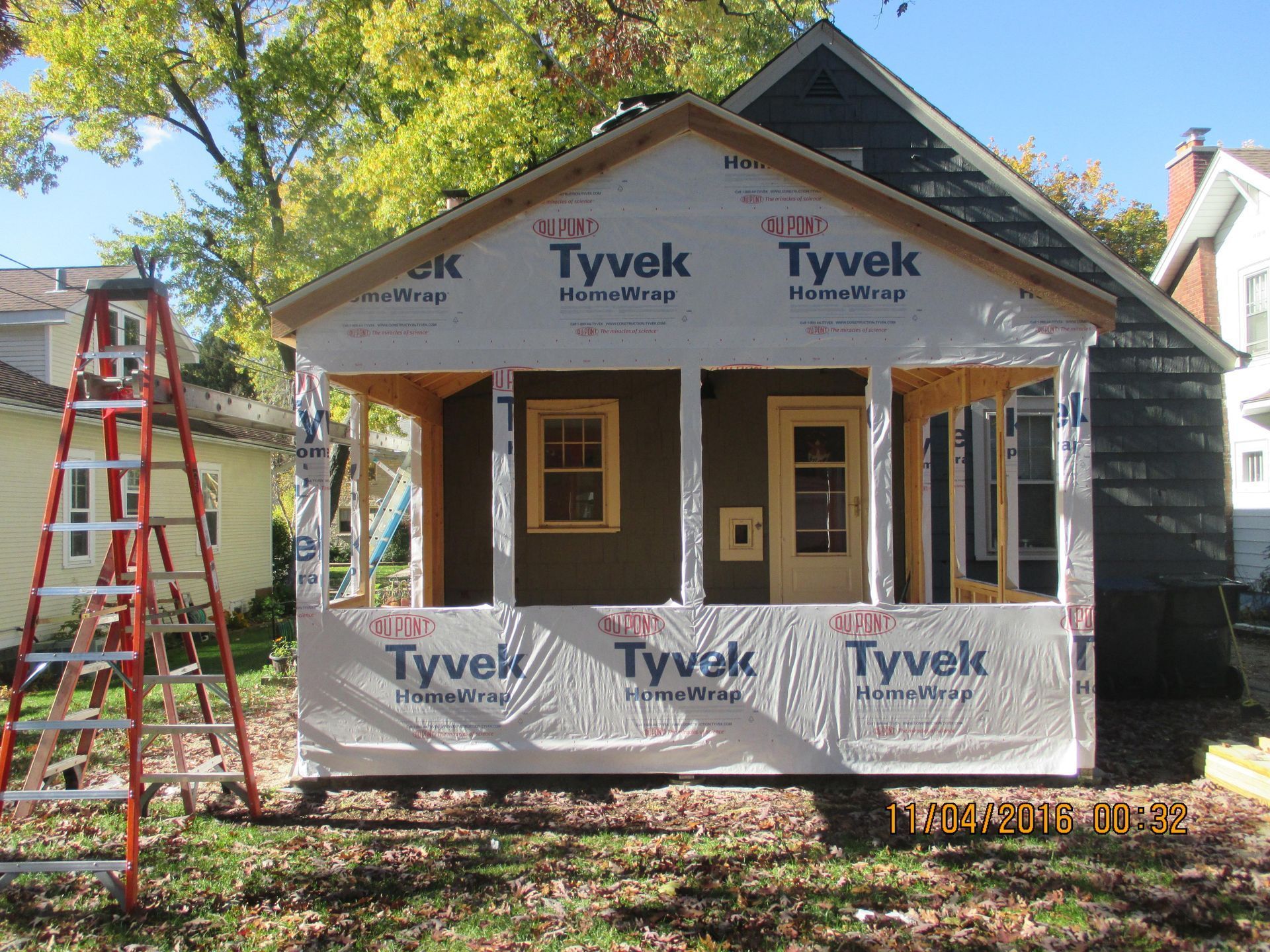 House under construction with porch, wrapped in Tyvek. Ladder to the left, blue sky, fall setting.