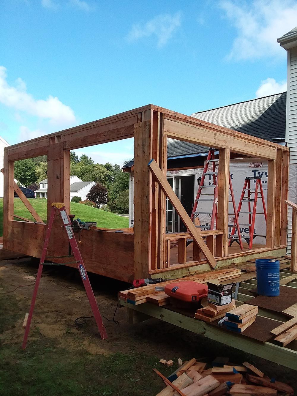 Construction of a wooden structure, possibly a deck or porch. Framing in progress, tools and materials scattered.