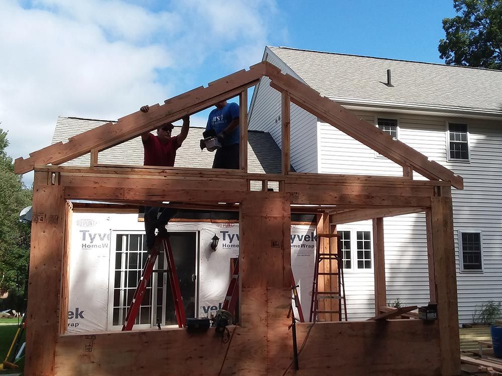 Construction of a wood-framed structure against a white house; workers installing roof beams.