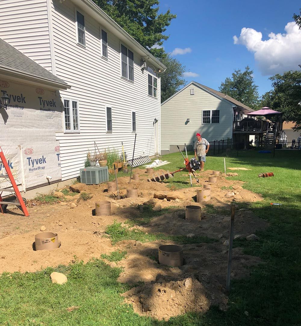 Construction site: House with exposed framing and holes dug in the yard; a worker operates equipment.