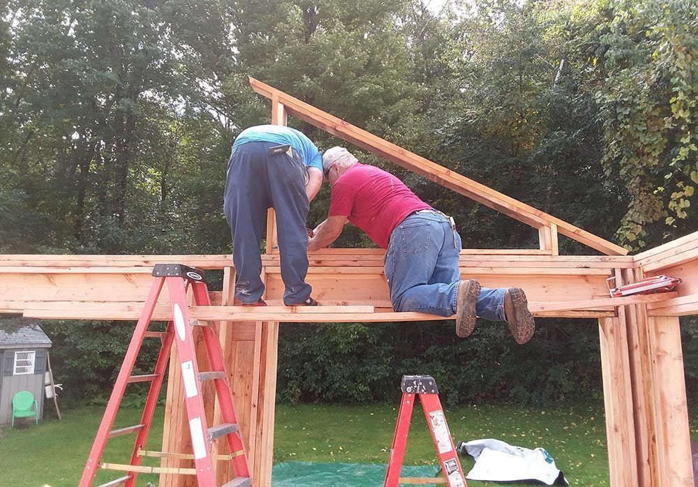 Two people working on a wooden roof structure, using ladders, outdoors.