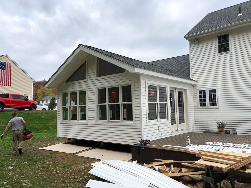 Sunroom addition attached to a white house with a deck. A worker carries tools across a lawn.