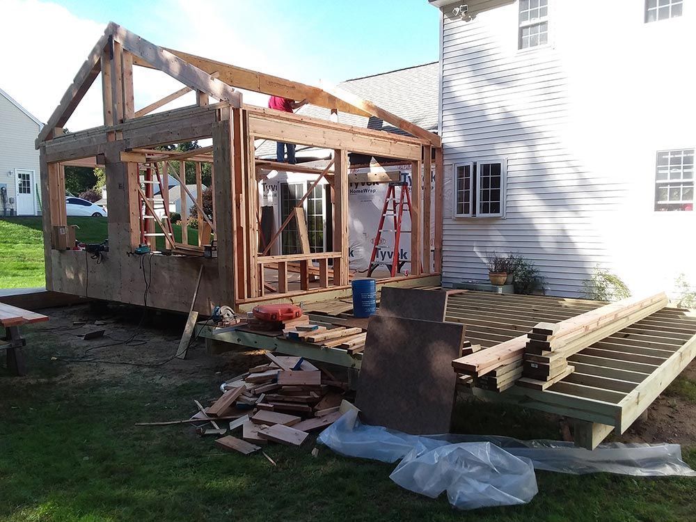 Construction of a wooden room addition attached to a white house, on a wooden deck, with scattered materials.
