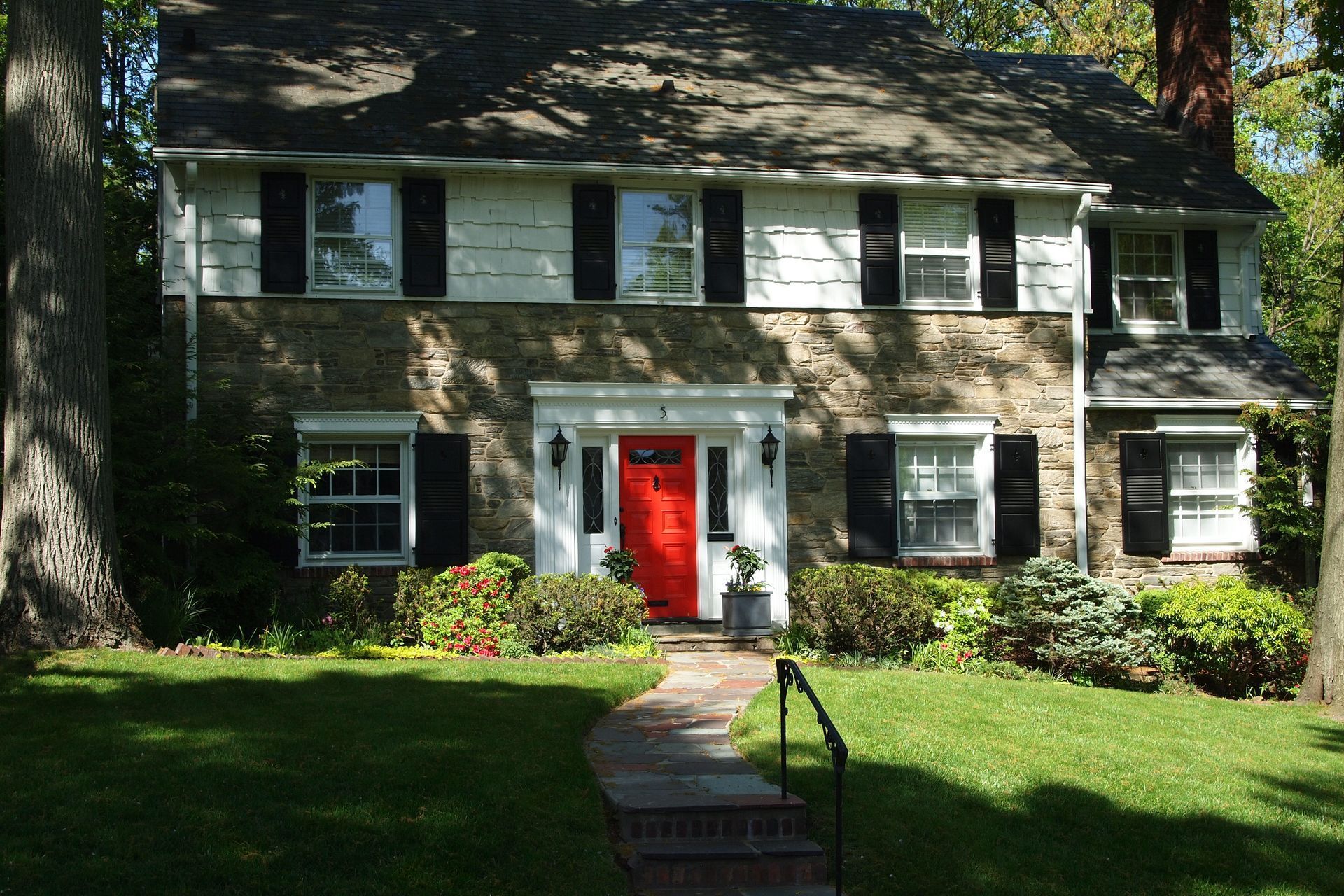 Two-story house with stone facade, black shutters, red front door, and a brick pathway.