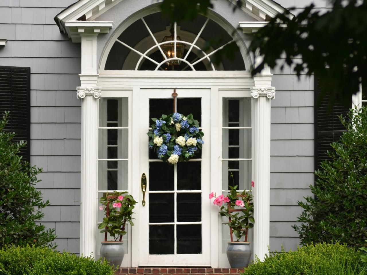 White front door with wreath, sidelights, and arched transom. Gray house with potted plants.
