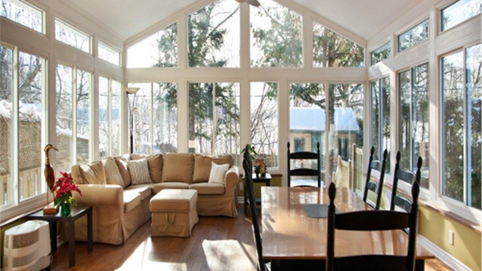 Sunroom with large windows, tan sectional sofa, and a dining table, overlooking a snowy outdoor scene.