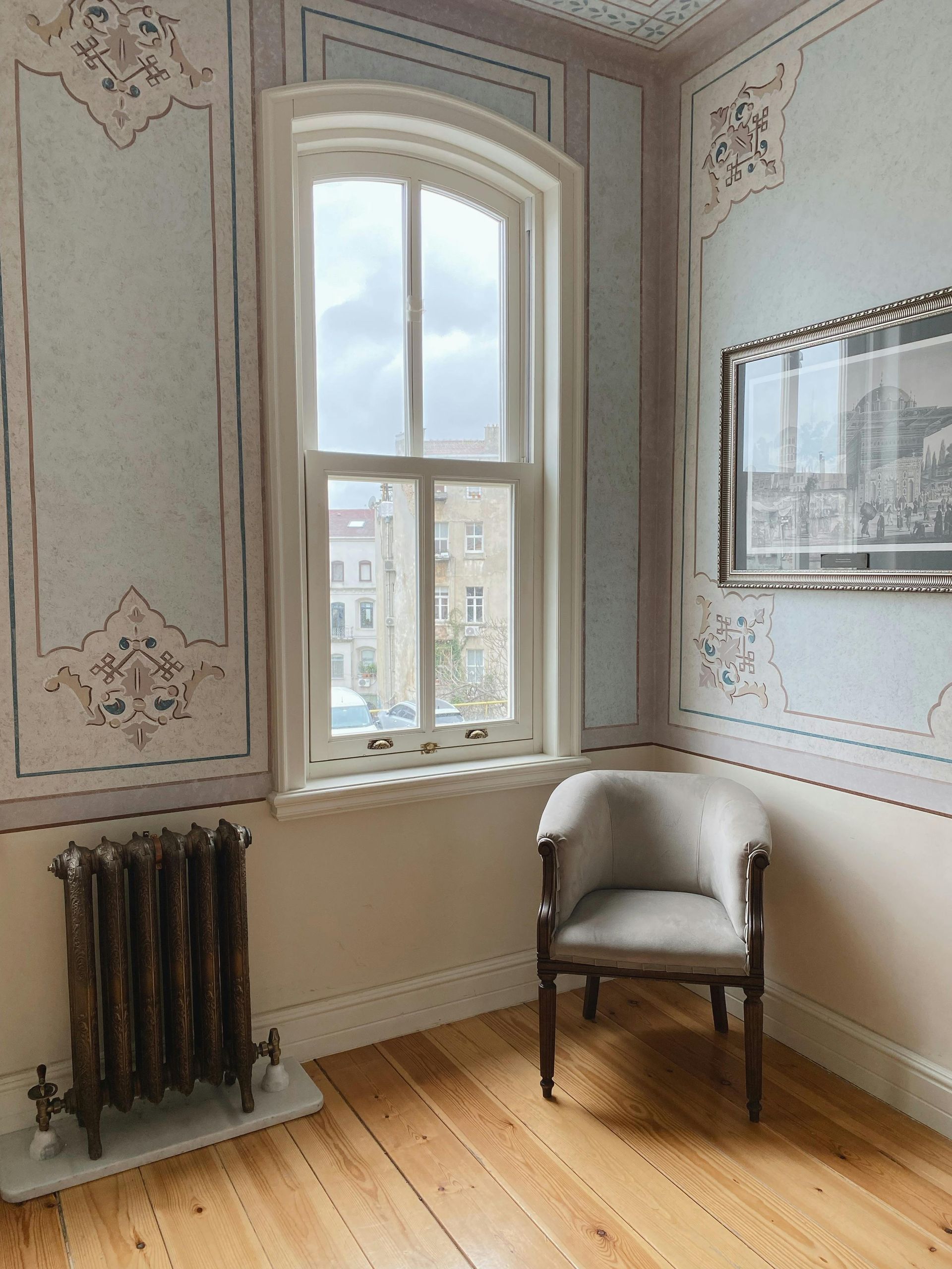 Room with ornate wallpaper, window, chair, radiator, and a framed picture.