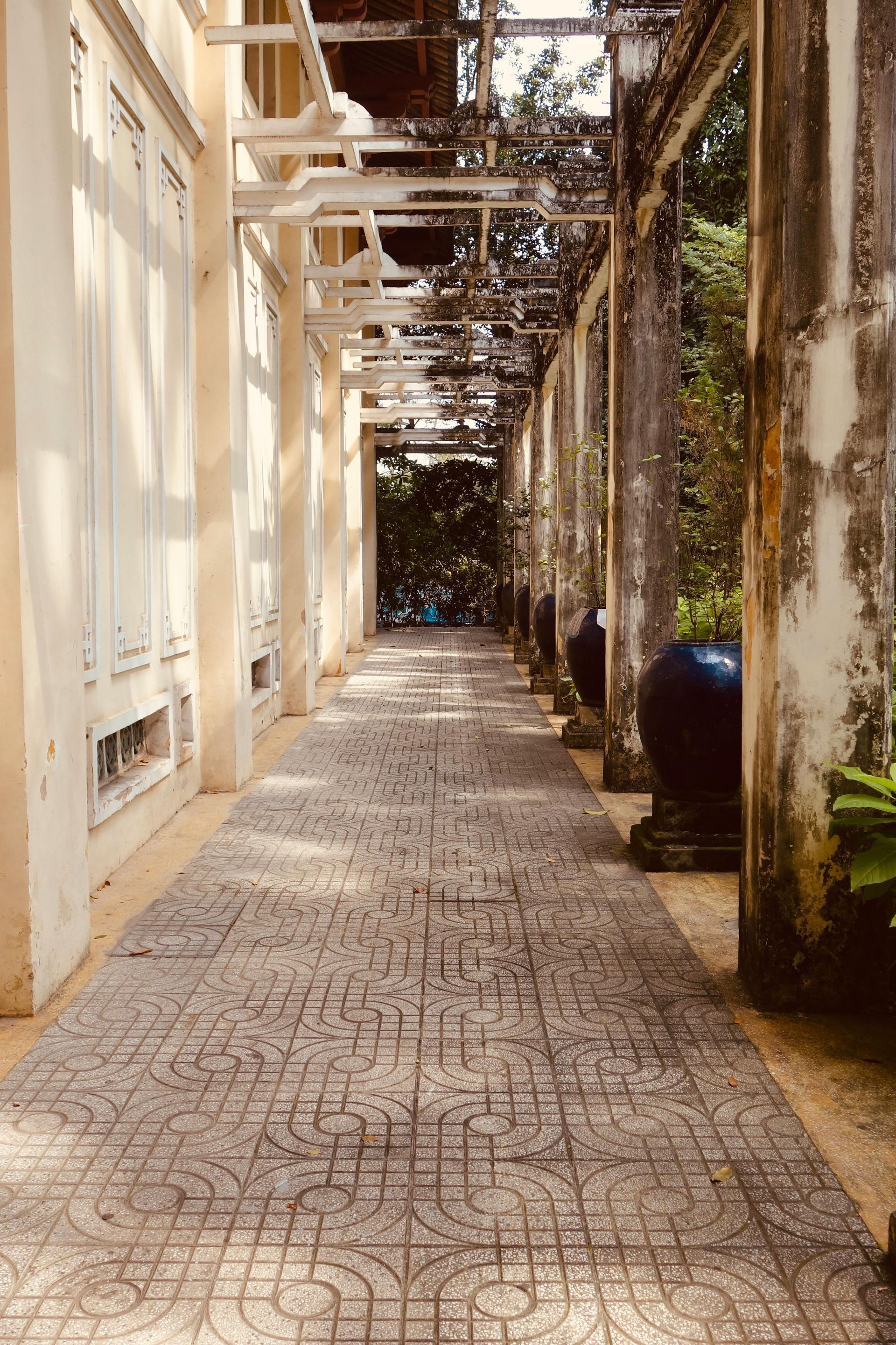 Covered walkway with stone floor, columns, and wooden trellis, leading towards greenery.