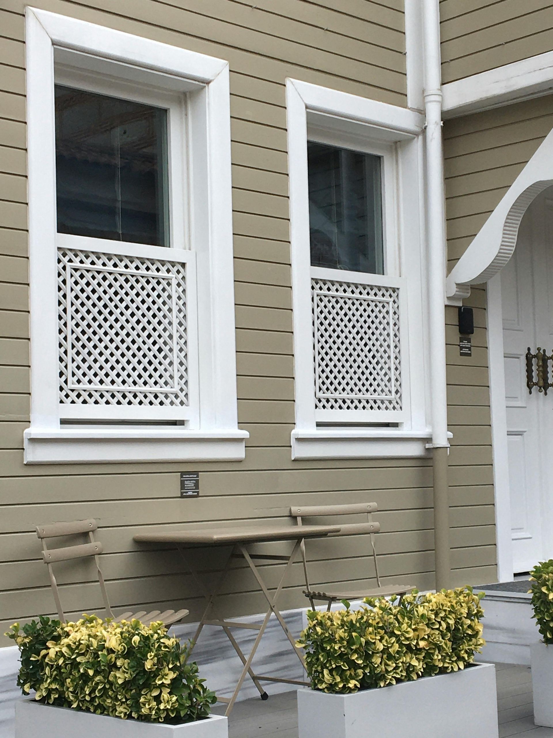 Two windows with white lattice panels on a light brown building exterior, a small table, chairs, and potted shrubs.