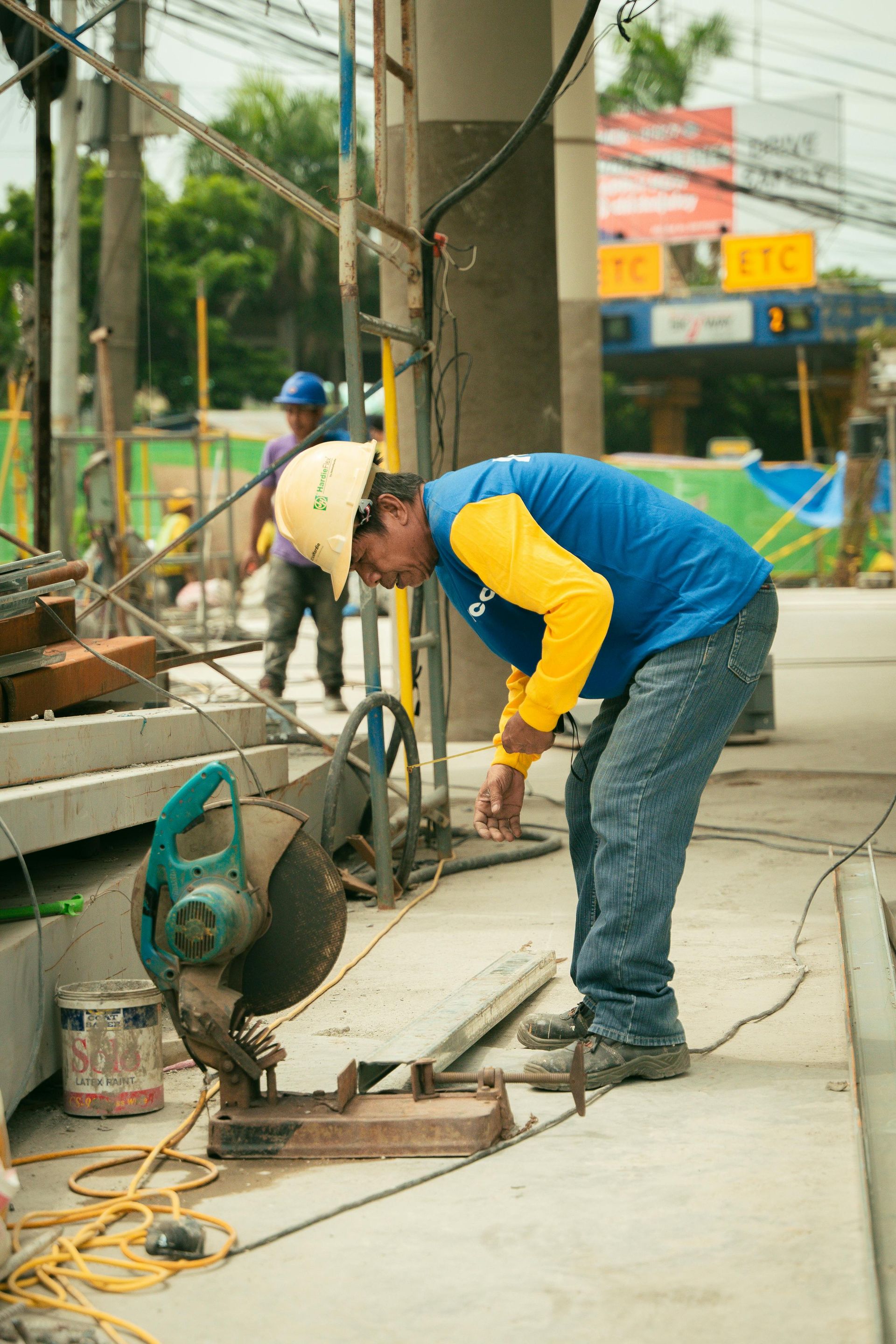 Construction worker in blue shirt and yellow hard hat cuts metal with a saw on a sidewalk.