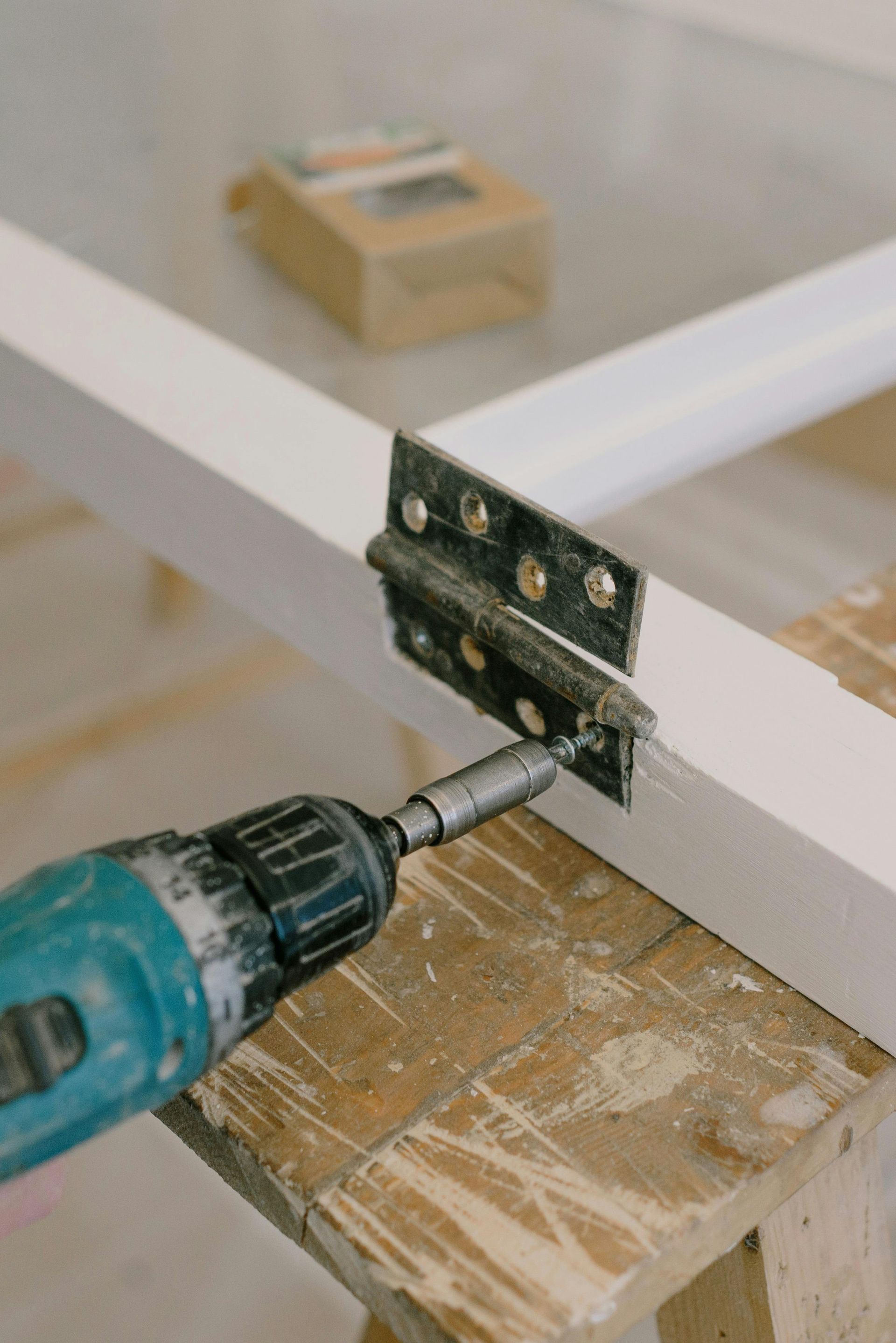 A person using a drill to attach a hinge to a white wooden frame on a workbench.