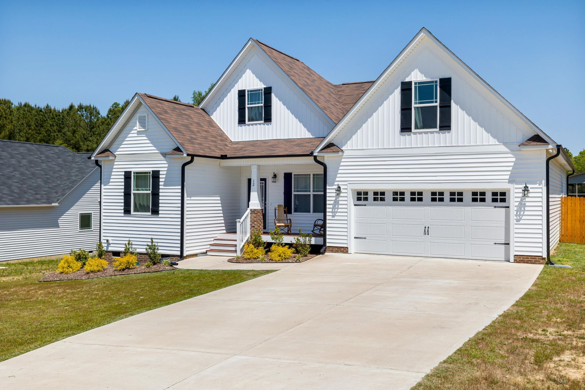 White two-story house with black shutters, brown roof, and concrete driveway on a sunny day.