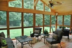 Sunroom with glass windows and outdoor seating, overlooking a green yard and trees.