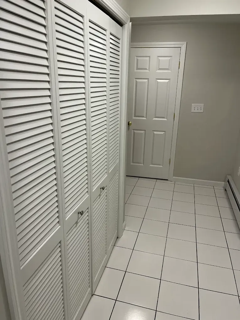 White louvered closet doors, white tile floor, a closed white door, and a light-colored wall.