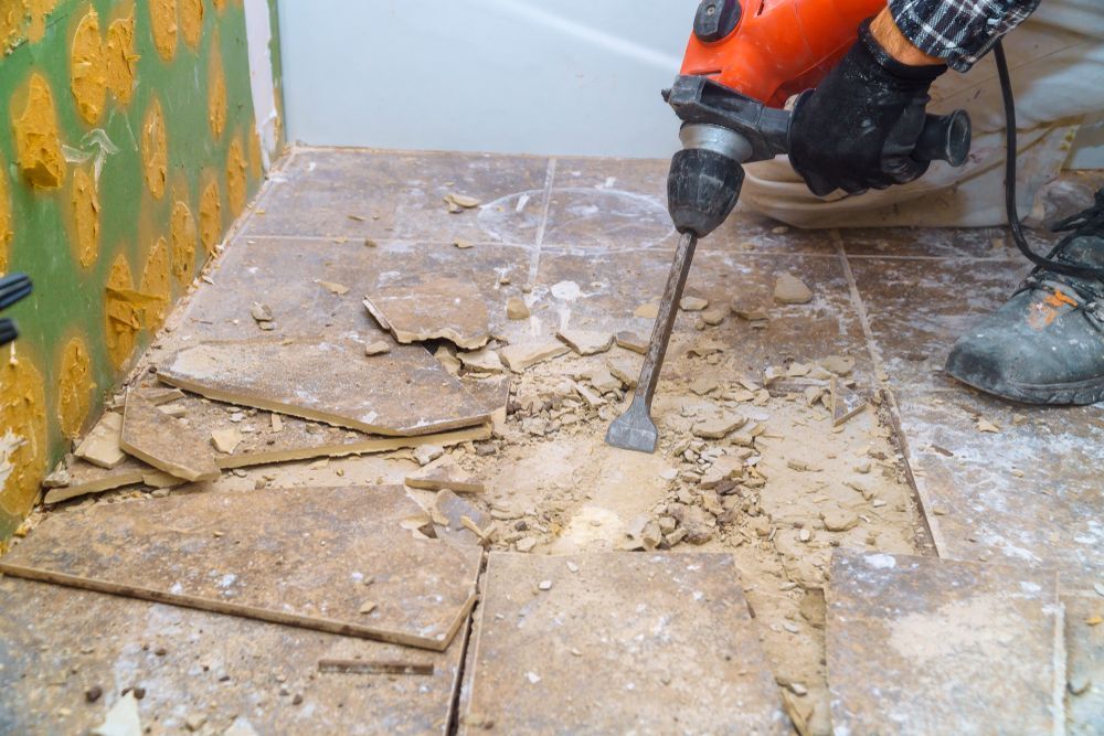A Person is Using a Hammer to Remove Tiles From the Floor — Shane Rice Builder & Renovation Specialist In Southside, QLD