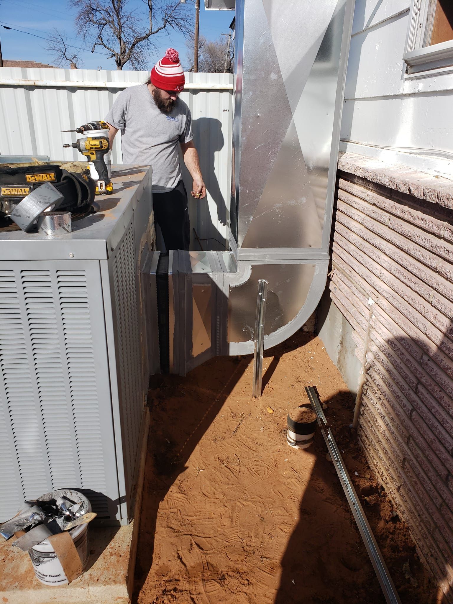 A man wearing a red hat is working on an air conditioner outside of a house.