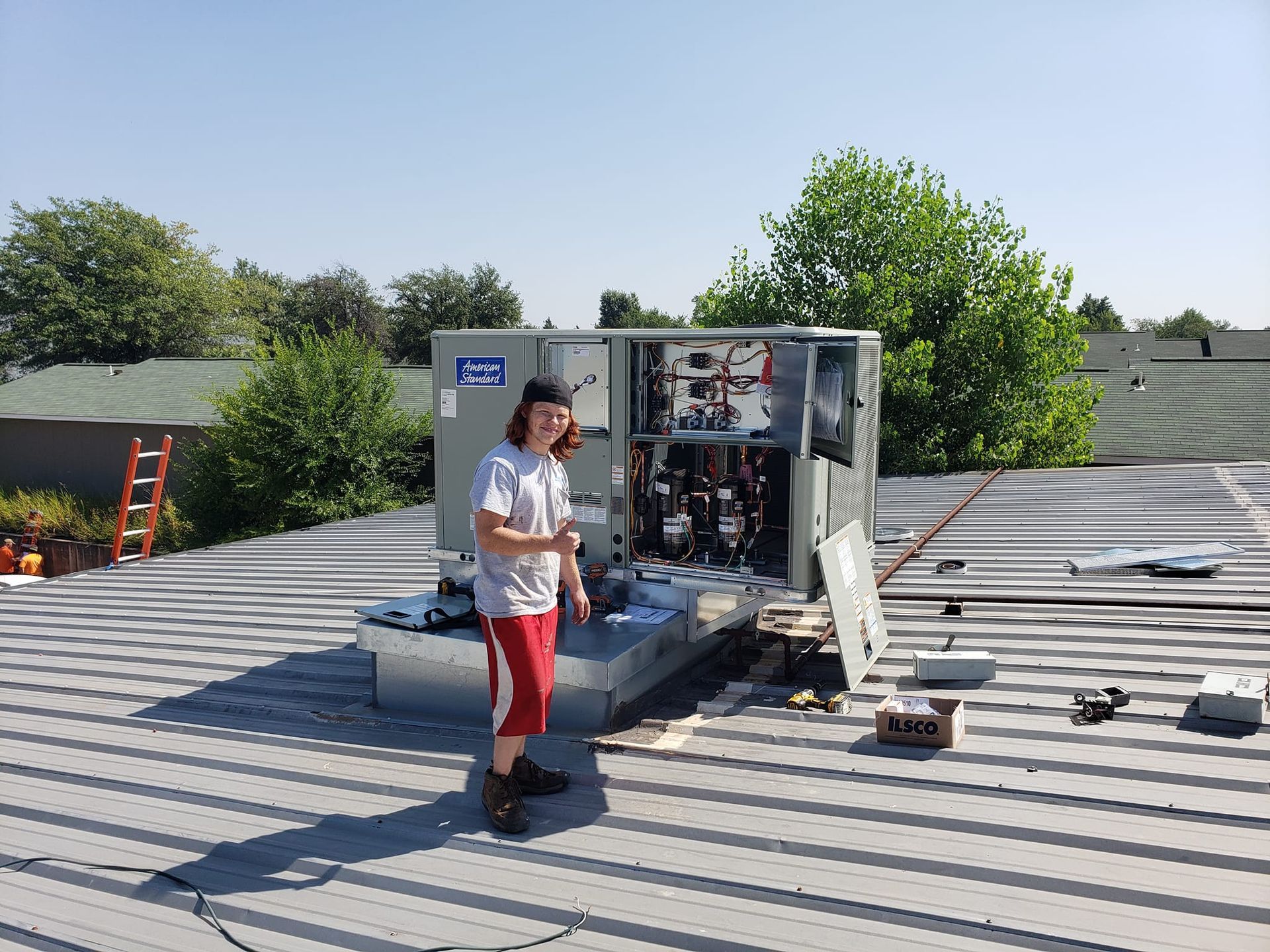 A man is standing on a roof working on an air conditioner.